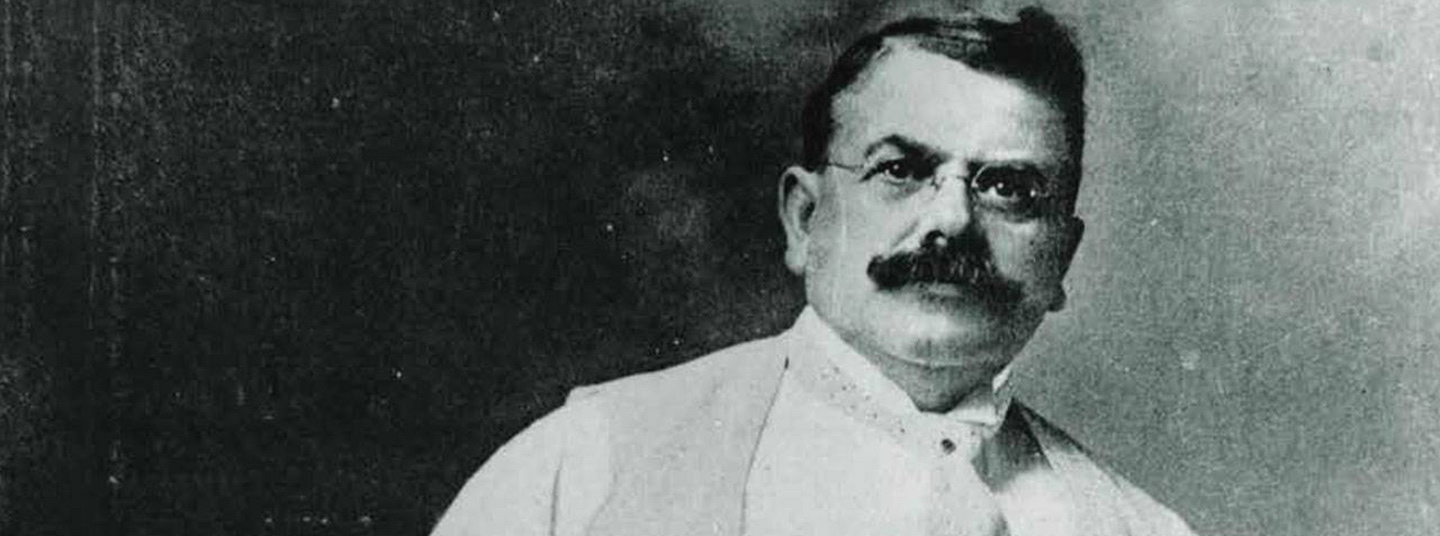 Photo en noir et blanc de Wallace C. Abbott.  Il a des cheveux noirs courts, une grosse moustache foncée et porte des lunettes, une chemise blanche, une veste de costume et une cravate.